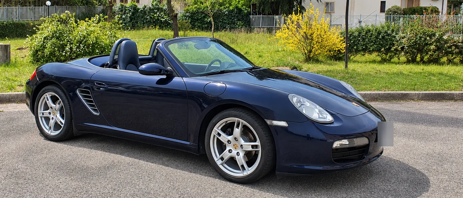 La Boxster blu scura con la capote aperta, ferma su un piazzale tra il verde in primavera
