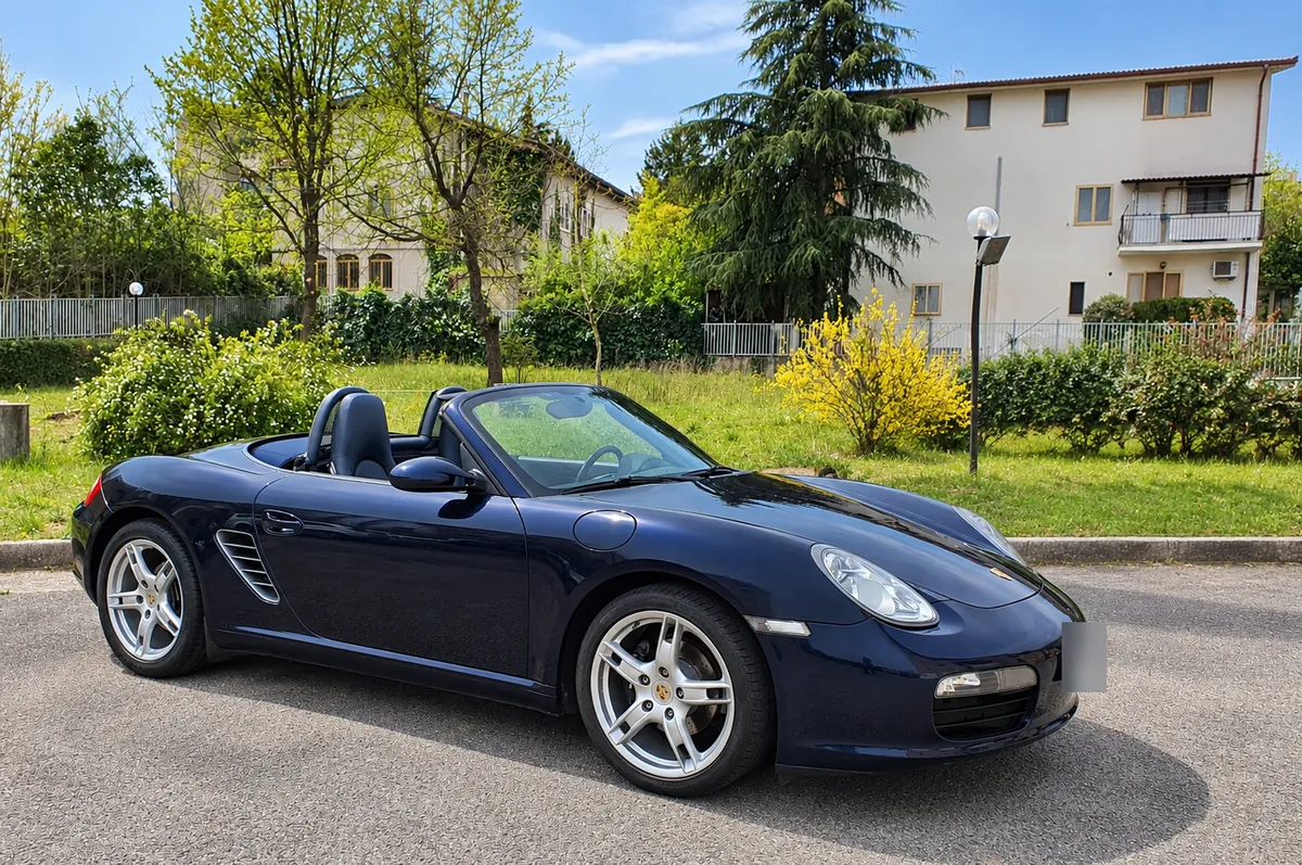 La Boxster blu scura con la capote aperta, ferma su un piazzale tra il verde in primavera