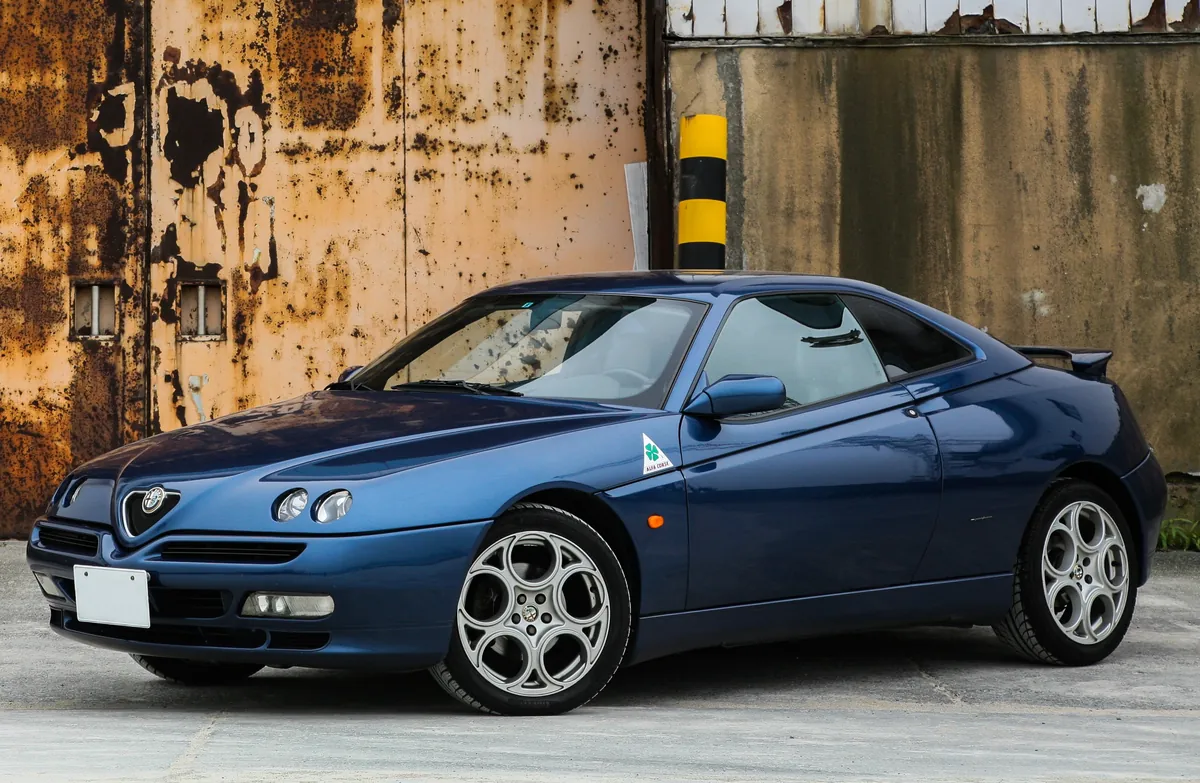 Alfa Romeo GTV blu davanti a un muro arrugginito